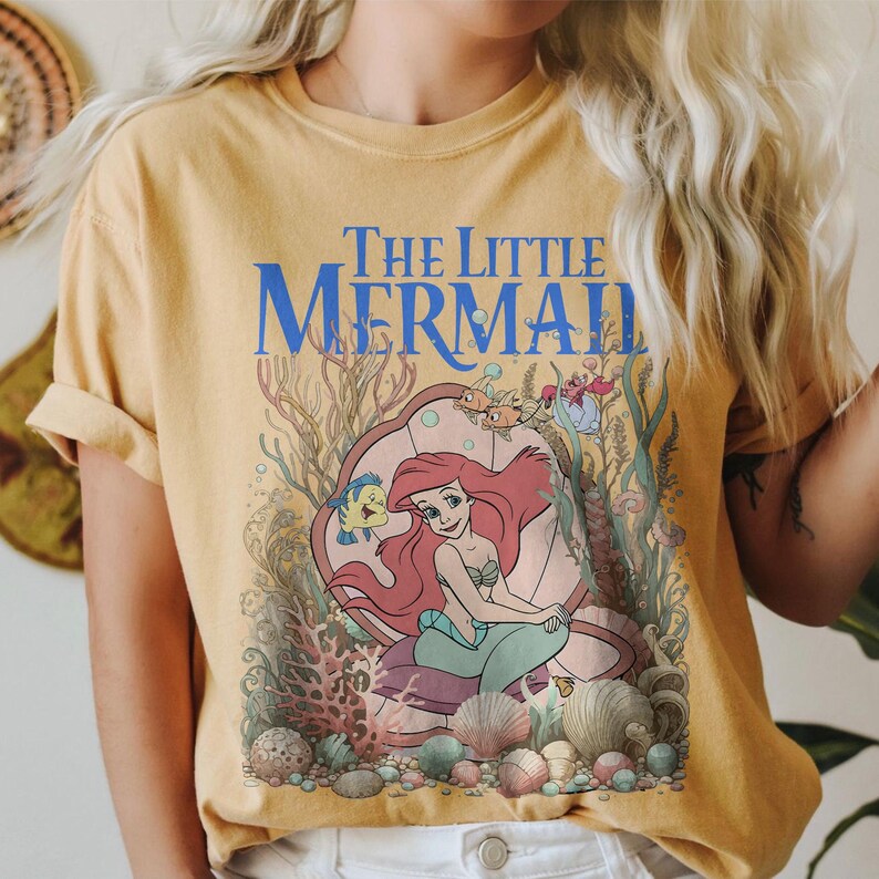 Op de afbeelding: Mosterdgeel T-shirt met een afbeelding van "De Kleine Zeemeermin". Het ontwerp bevat Ariel, Botje en Sebastiaan, omringd door koraal en schelpen. De titel "The Little Mermaid" staat in blauwe letters.