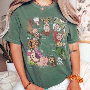 May include: Green t-shirt featuring a colorful graphic of characters from the animated film Toy Story. The design includes Woody, Buzz Lightyear, Jessie, and other characters. The shirt is a casual style, suitable for everyday wear.