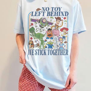 May include: Light blue t-shirt featuring a colorful graphic of Toy Story characters, including Woody, Buzz Lightyear, and Rex. The shirt has the text "NO TOY LEFT BEHIND WE STICK TOGETHER" in a retro style.