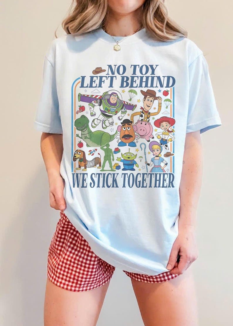 May include: Light blue t-shirt featuring a colorful graphic of Toy Story characters, including Woody, Buzz Lightyear, and Rex. The shirt has the text "NO TOY LEFT BEHIND WE STICK TOGETHER" in a retro style.