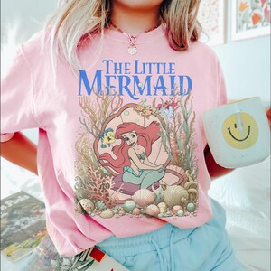 Op de afbeelding: Zachtroze T-shirt met de blauwe tekst "The Little Mermaid" boven een illustratie van Ariel, Botje en Sebastiaan. De afbeelding bevat schelpen, koraal en ander zeeleven. Een witte mok met een gele smiley wordt in de afbeelding vastgehouden.