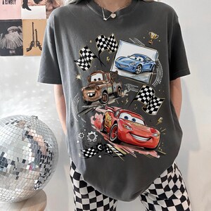 Op de afbeelding: Donkergrijs t-shirt met een kleurrijke afbeelding van geanimeerde raceauto's, waaronder een rode auto met nummer 95, een bruine takelwagen en een blauwe sportwagen. Geblokte vlaggen en een trofee zijn ook afgebeeld.