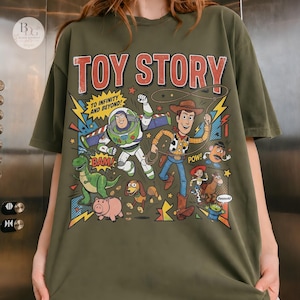 May include: Olive green t-shirt featuring a colorful graphic from the movie "Toy Story." The design includes the title "TOY STORY" in red, along with characters Buzz Lightyear, Woody, and others. Text includes "TO INFINITY AND BEYOND!" and sound effects.