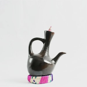 May include: A dark brown, polished ceramic coffee pot with a long, slender neck and a curved handle. The base is wrapped in a decorative band of pink, white, and blue woven fibers. A small, pointed, multi-colored conical stopper sits in the opening.