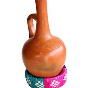 May include: A terracotta pitcher with a rounded body, a tall narrow neck, and a single handle, resting inside a circular woven base featuring alternating panels of bright turquoise and magenta with a white geometric diamond pattern woven throughout.