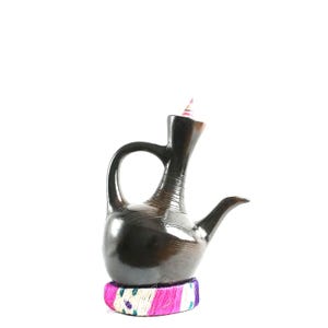 May include: A dark brown, polished ceramic coffee pot with a long, slender neck and a curved handle. The base is wrapped in a decorative band of bright pink, white, and teal fibers. A small, pointed, multi-colored stopper sits in the opening.