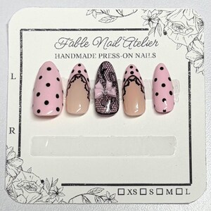 May include: A set of five almond-shaped press-on nails on a Fable Nail Atelier display card. The design features light pink nails with black polka dots, nails with a nude base and black lace trim, and a central black lace nail with a pink bow.