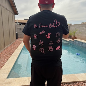May include: Black t-shirt with pink text and graphics. The text reads "My Forever Girl" with a heart symbol. Below the text are various pink illustrations. A red baseball cap is worn.