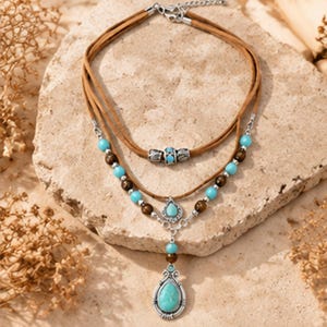 May include: A multi-strand necklace featuring brown suede cords and silver-toned metal accents. The design incorporates round turquoise and dark brown beads, with a central silver-toned pendant holding a teardrop-shaped turquoise stone.