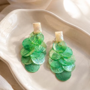 May include: A pair of dangle earrings featuring a rectangular cream-colored top stud and a cascading cluster of circular, translucent green shell discs. The discs are layered in a tiered arrangement, creating a textured, shimmering effect.