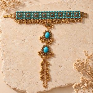 May include: A gold-toned metal hand chain featuring a wide cuff adorned with small turquoise beads in a square pattern and dangling gold spheres. A central vertical chain connects to two oval turquoise cabochons and ends in a finger loop.