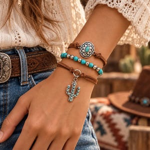 Può includere: Un set di bracciali a più fili con cordini in pelle marrone, perline color turchese, accenti metallici color argento, un ciondolo centrale ovale con inserto turchese e un ciondolo a forma di cactus color argento con dettagli turchesi.