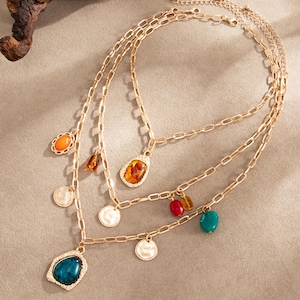 May include: A multi-layered gold-tone chain necklace featuring various charms, including smooth orange, amber, red, and teal oval stones, alongside several small, textured, round gold-tone metal discs hanging from the links.