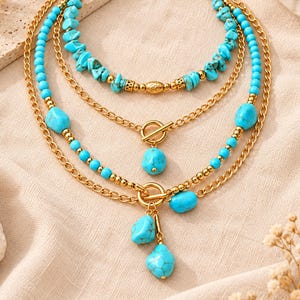 May include: A multi-layered necklace featuring gold-tone chains interspersed with bright turquoise stone beads. The design includes two toggle clasps, one with a single turquoise pendant and the other with two dangling turquoise stone charms.