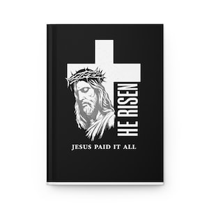 May include: A black hardcover journal featuring a white graphic of a cross and a portrait of Jesus wearing a crown of thorns. The text HE RISEN is printed vertically next to the cross, and the text JESUS PAID IT ALL is printed below the portrait.