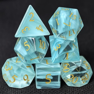 Könnte beinhalten: Ein Satz von sieben Würfeln in verschiedenen Formen. Die Würfel sind hellblau transparent mit weißen Marmorierungen und goldenen Zahlen. Das Set enthält einen d4, d6, d8, d10, d12, d20 und einen Prozentwürfel. Für Tabletop-Spiele.