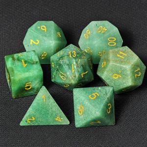 Könnte beinhalten: Set aus sieben grünen Edelsteinwürfeln mit goldfarbenen Zahlen. Die Würfel umfassen einen d4, d6, d8, d10, d12, d20 und einen Prozentwürfel. Die Würfel liegen auf einer dunklen Oberfläche.