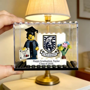 May include: A Lego graduation display with a minifigure in a cap and gown holding a certificate. The display includes a university crest, flowers, and the text "Happy Graduation, Taylor Class of 2026". The display is encased in a clear box.