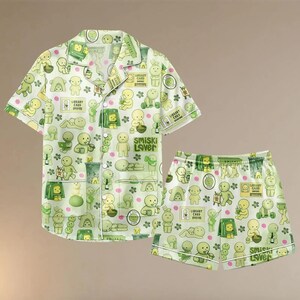 Puede incluir: Conjunto de pijama verde claro con un patr&oacute;n repetido de figuras de dibujos animados, flores y dise&ntilde;os de tarjetas de biblioteca. El conjunto incluye una camisa con botones y pantalones cortos a juego. El texto "Smiski Lover" est&aacute; impreso.