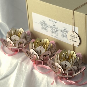 Könnte beinhalten: Drei lasergeschnittene Lotusblumen aus Holz mit rosa Blütenblättern und goldfarbenen Zentren, jede mit einem rosa Band. Die Blumen befinden sich vor einer Pappschachtel mit einem Etikett mit der Aufschrift "Red Stem" und "The Lotus".