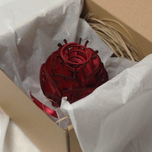 May include: A red, laser-cut wooden rose-shaped ornament. The item is presented in a cardboard box lined with white tissue paper. A red ribbon and dried grass are also visible.