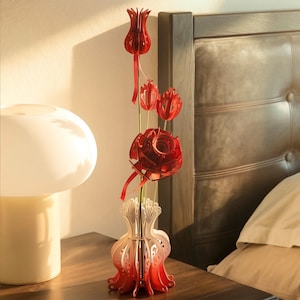May include: A decorative sculpture featuring three red, layered, tulip-shaped flowers on thin stems, emerging from a white and red, laser-cut, pomegranate-shaped base. The structure is composed of interlocking pieces with intricate, cut-out patterns.
