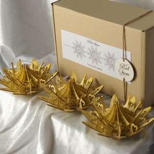 May include: Three laser-cut wooden sunflower decorations in a warm yellow hue are displayed on a white surface. A cardboard box with a label that reads "Red Stem" and "THE SUNFLOWER" is in the background. A wooden tag with the words "Red Stem" hangs from the box.