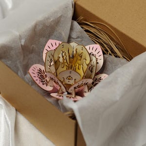 Può includere: Un fiore di loto in legno tagliato al laser in una scatola di cartone, con carta velina. Il fiore presenta petali rosa e crema con intricati disegni ritagliati. La rafia decorativa aggiunge un tocco naturale. Un'idea regalo unica.