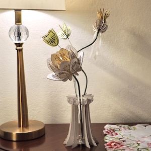 May include: A three-dimensional laser-cut wooden sculpture of a flower arrangement in a vase. The vase is shaped like a classical column. The flowers feature layered petals in shades of white and pale yellow, with thin stems and a seed pod.