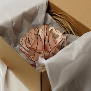 Può includere: Una scultura floreale in legno rosa, tagliata al laser, in una scatola di cartone con carta velina bianca. Il fiore presenta dettagli intricati ed è parzialmente assemblato. La scatola è aperta.