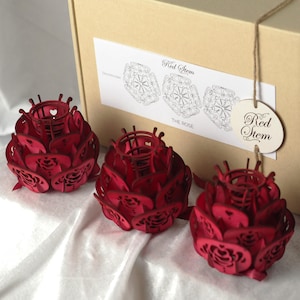 Puede incluir: Tres candelabros rojos de metal con forma de rosa. Cada uno tiene una parte superior circular con un recorte en forma de corazón y diseños intrincados de pétalos. Los candelabros se exhiben sobre una superficie blanca con una caja beige en el fondo. La caja tiene una etiqueta que dice "Red Stem".