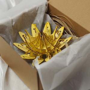 May include: A yellow, laser-cut wooden decorative object, resembling a stylized flower or crown, is nestled in a cardboard box lined with white tissue paper. The object has intricate cut-out heart and dot patterns.