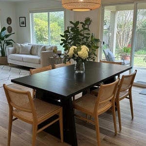 May include: A dining room set with a dark wood table and six chairs with woven backs. A large woven pendant light hangs above the table. The room has hardwood floors, a white sofa, and large windows overlooking a backyard.