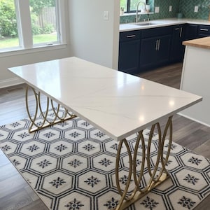 May include: A rectangular white marble-look dining table with gold-colored, curved metal legs. The table sits on a patterned rug with a black and white geometric design. The room has large windows and a modern kitchen.