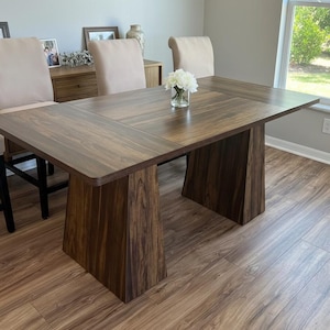 May include: A dark brown wooden dining table with a rectangular top and two trapezoidal supports. The table is surrounded by beige upholstered chairs. A vase of white flowers sits on the table. The room has a large window and hardwood floors.