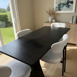 Puede incluir: Un juego de comedor moderno con una mesa de madera negra y cuatro sillas tapizadas blancas. La mesa tiene un acabado oscuro y un diseño de base único. Las sillas tienen respaldos curvos y patas de metal negro. La habitación tiene paredes claras y un armario de madera.