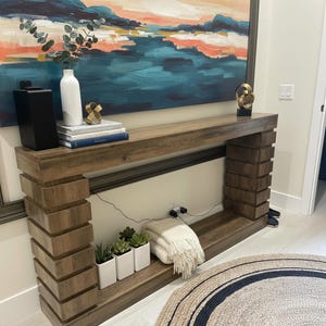 May include: A wooden console table with a geometric design, featuring a lower shelf. The table is adorned with decorative items, including a white vase with greenery, books, and small potted plants. A round rug with a blue border is in the foreground.