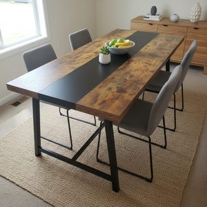 May include: A rectangular dining table with a distressed wood finish and a black center stripe. The table has black metal legs and is surrounded by four gray upholstered chairs. A bowl of fruit and a small potted plant sit on the table.