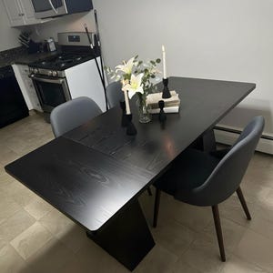 May include: A black dining table with a geometric design, paired with gray chairs. The table is set with white flowers in a glass vase, black candle holders, and lit candles. The table measures approximately 72 inches long.