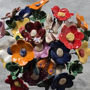 Könnte beinhalten: Ein Blumenstrauß aus handgefertigten Keramikblumen und Schmetterlingen in verschiedenen Farben wie Rot, Orange, Gelb, Blau, Lila und Weiß. Jede Blume hat eine strukturierte Mitte und grüne Keramikblätter sind im gesamten Arrangement verteilt.