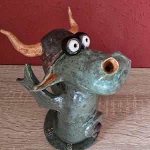 Op de afbeelding: Een keramieken sculptuur van een groene draak met een textuurafwerking, voorzien van een donkerbruine helm met beige hoorns, grote witte ogen met zwarte pupillen en een holle snuit. De draak heeft kleine vleugels en staat op een ronde voet.