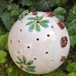 Könnte beinhalten: Ein kugelförmiges weißes Keramikobjekt mit zahlreichen kleinen kreisförmigen Perforationen auf der Oberfläche. Das Stück ist mit grün bemalten Blumenmotiven und mehreren kleinen, braunen, texturierten Igel-Figuren an der Außenseite verziert.