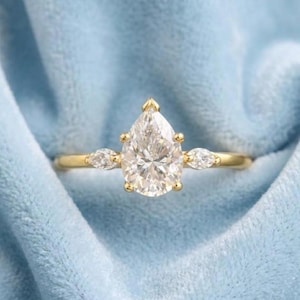 Op de afbeelding: Een gouden ring met een grote, peervormige diamant als middelpunt. Twee kleinere, marquise-geslepen diamanten flankeren de hoofdsteen. De ring is geplaatst op een zachte, lichtblauwe achtergrond.