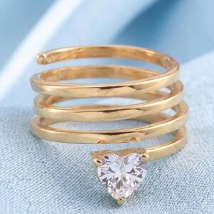 May include: A gold-coloured spiral ring featuring a heart-shaped, clear gemstone. The ring coils around the finger several times, creating a unique design. The gemstone is set in a gold-coloured bezel.