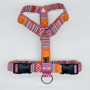 May include: A colorful dog harness with a woven pattern in shades of orange, pink, purple, and yellow. The harness has black plastic buckles and a metal ring for leash attachment. A small brown leather patch is centered on the harness.