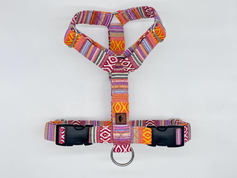 May include: A colorful dog harness with a woven pattern in shades of orange, pink, purple, and yellow. The harness has black plastic buckles and a metal ring for leash attachment. A small brown leather patch is centered on the harness.