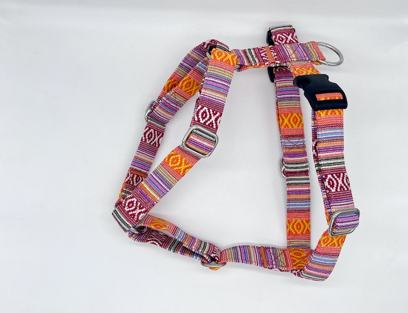 May include: A colorful dog harness with a woven pattern in shades of red, orange, purple, and yellow. The harness features adjustable straps, metal hardware, and a black plastic buckle. The design includes geometric shapes and stripes.