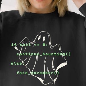 Pode incluir: Uma camisola preta de manga comprida com um gráfico de fantasma em linhas brancas. O design inclui texto de código informático em verde que diz: if soul != 0: continue_haunting() else: face_november(). A peça tem gola redonda.