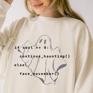 May include: An ivory long-sleeve sweatshirt featuring a dark gray graphic of a ghost and black computer code text that reads: if soul == 0: continue_haunting() else: face_november(). The sweatshirt has a crew neckline and ribbed cuffs.