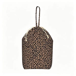 May include: A drawstring bag featuring a tan and black leopard print pattern. The bag includes a single top handle made of the same leopard print fabric and white cord drawstrings with small tassels at the ends for secure closure.